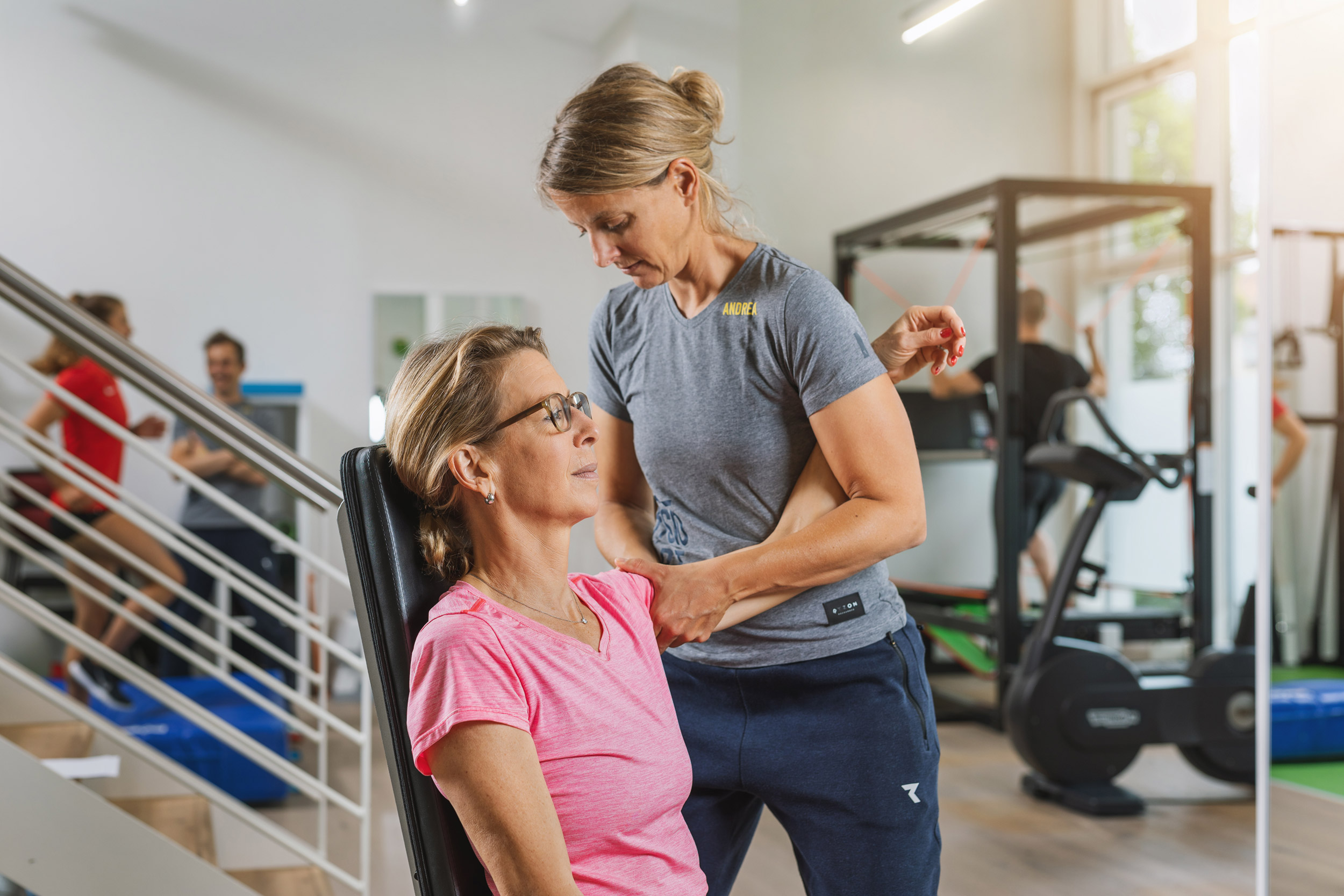 Physiotherapeutin behandelt eine Patientin an der Schulter um ihre Beweglichkeit zu verbessern. Im Hintergrund sieht man den Trainingsraum, welcher von weiteren Personen genutzt wird.