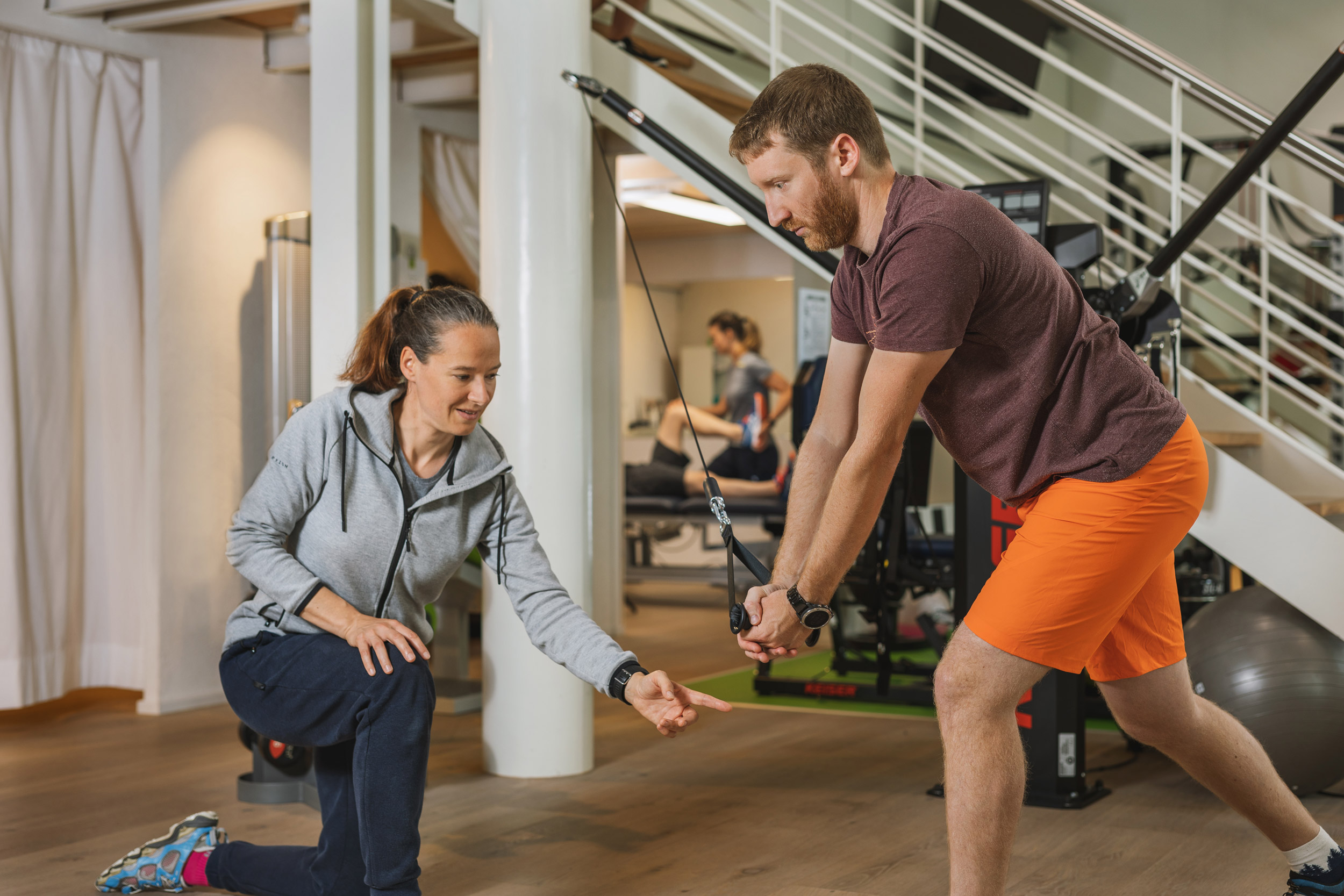 Eine Physiotherapeutin instruiert eine Trainingsübung und coacht ihren Kunden. Im Hintergrund sieht man in einen Behandlungsraum, wo eine weitere Physiotherapeutin arbeitet.